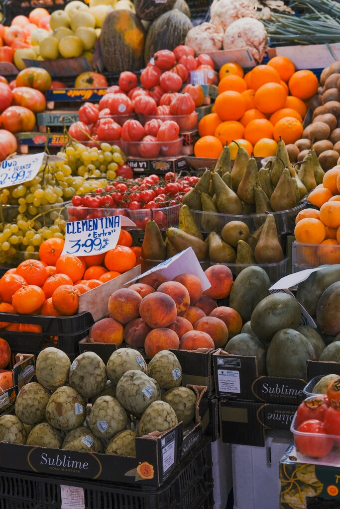 Woman photographing fresh produce at a Mediterranean market with a sketch-filled tote bag